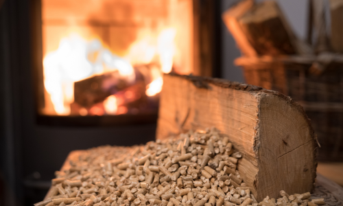 A split log of firewood and pile of pellets in front of a lit pellet stove